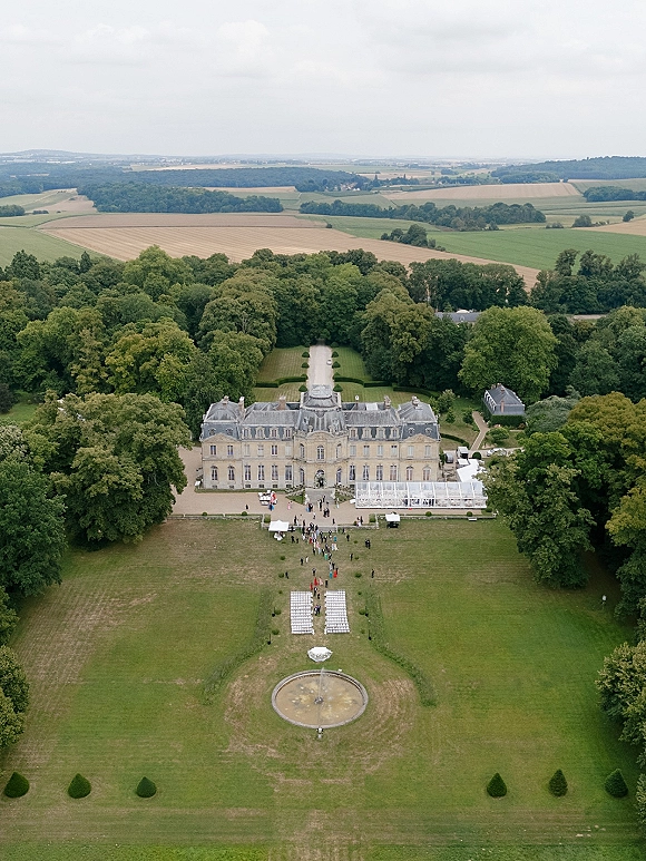 Estate wedding venue with a clear reception tent and outdoor ceremony chairs beside a fountain on manicured lawns, ringed by trees