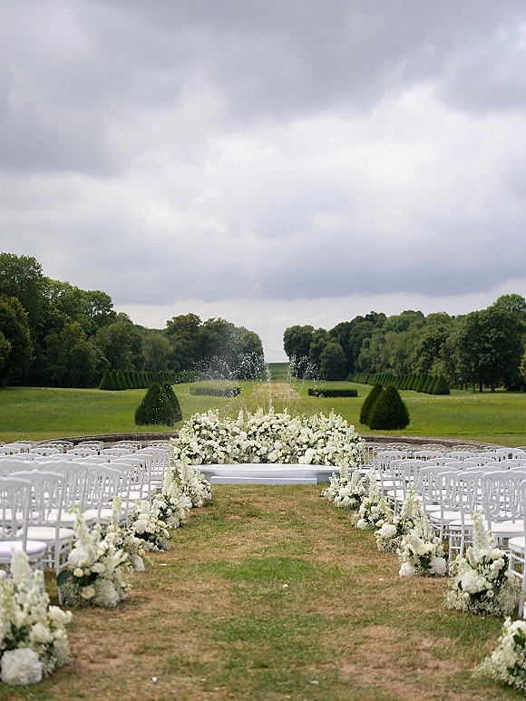 Ceremony setup for an outdoor wedding ceremony with white aisle florals and altar arrangement, white chairs on a lawn with garden vista