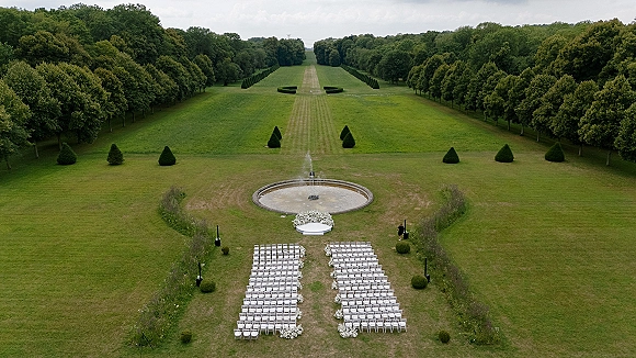 Outdoor ceremony setup with white folding chairs and aisle florals leading to a fountain on a manicured, tree-lined lawn under overcast sky