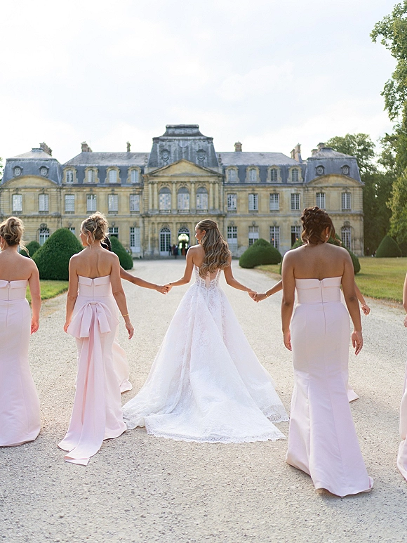Bride with bridesmaids walking away holding hands, lace gown train flowing beside strapless blush dresses on a chateau driveway
