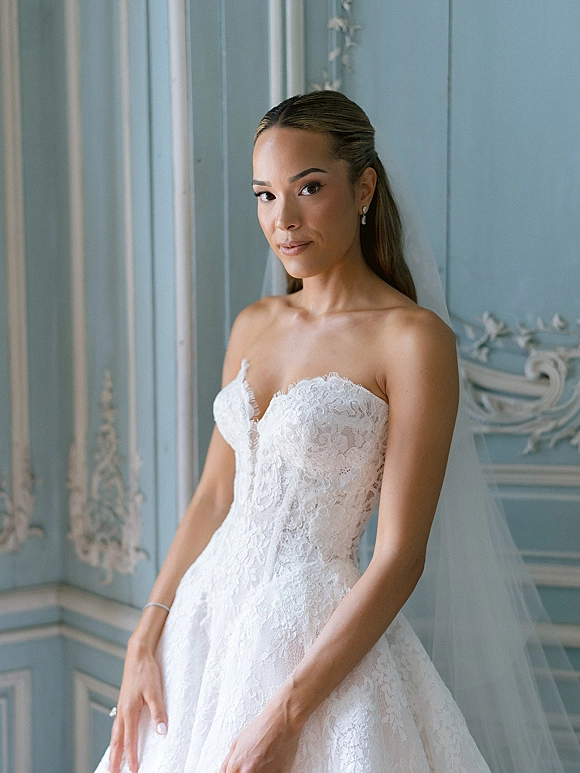 Bridal portrait of a bride in a strapless lace wedding dress with tulle veil and drop earrings, posed against ornate blue wall paneling
