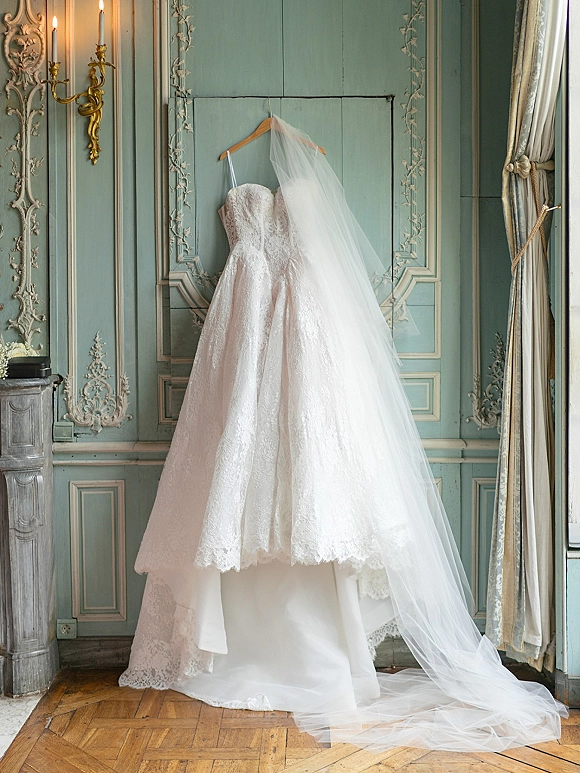 Wedding dress with lace wedding dress detailing hangs on a wooden hanger, long tulle veil draped against an ornate paneled wall in window light