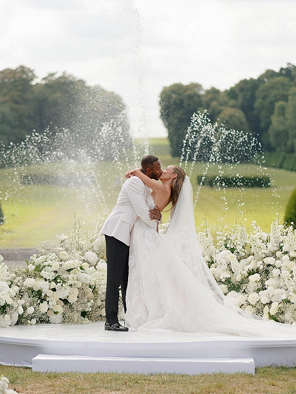 Wedding kiss portrait of bride and groom kissing, her long veil flowing as they dip before a garden fountain with white florals