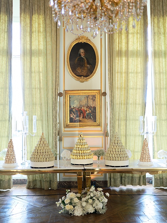 Dessert table with wedding dessert table macaron towers, glass candelabras, white florals, and a chandelier before tall curtained windows