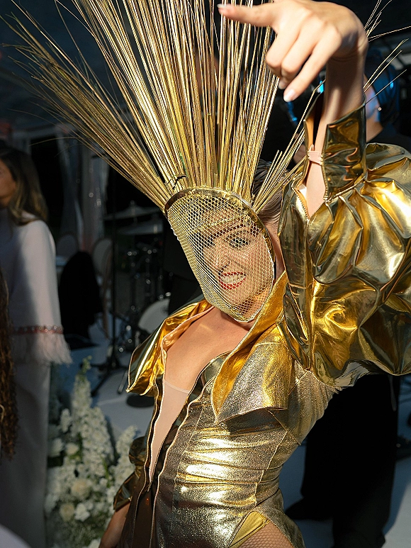 Wedding guest fashion in a metallic gold outfit with dramatic headpiece and mesh face veil, red lipstick, near a crowd and floral display