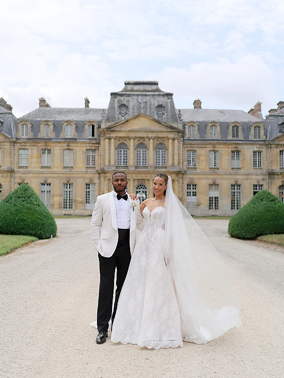 Couple portrait of bride in lace ballgown with cathedral veil and groom in white tuxedo on a gravel drive before a historic mansion