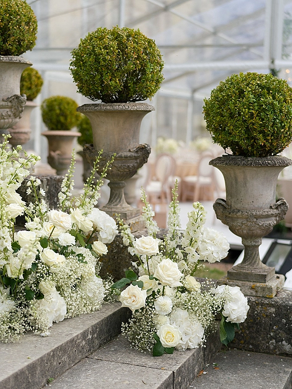 Wedding floral decor with white wedding flowers cascading along stone steps, featuring roses, hydrangeas, baby's breath, and urn planters by a glass tent