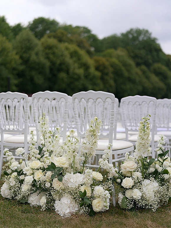 Ceremony aisle decor with outdoor ceremony chairs, low white rose and hydrangea florals with greenery lining a grassy lawn under trees