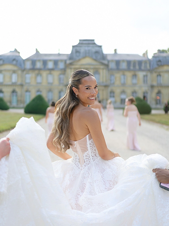 Bride portrait in a strapless lace wedding dress, looking back and holding her train on a gravel drive beside a historic estate facade