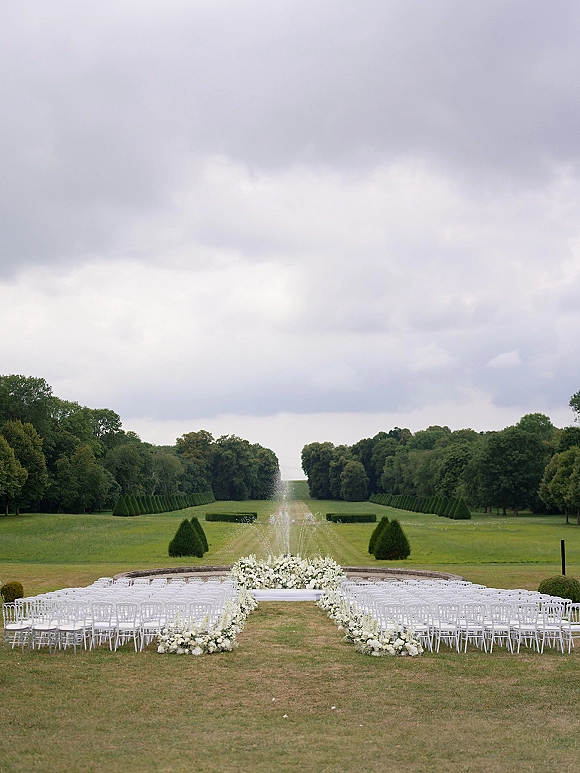 Ceremony setup with white chair ceremony seating and aisle florals leading to a floral altar on a raised stage in a formal garden with fountain