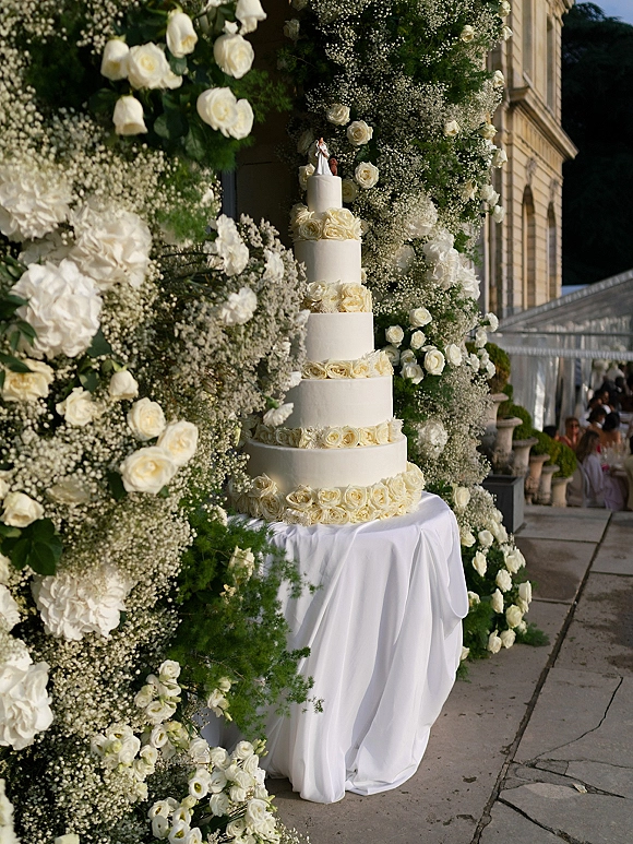 Wedding cake with five tier white buttercream, sugar roses and topper figurines, framed by a white rose garland and floral wall on a stone terrace