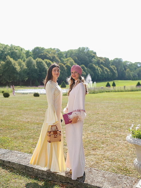 Wedding guest portrait of two women in long sleeve maxi dresses, one with a turban headwrap, posing by a fountain on a manicured lawn