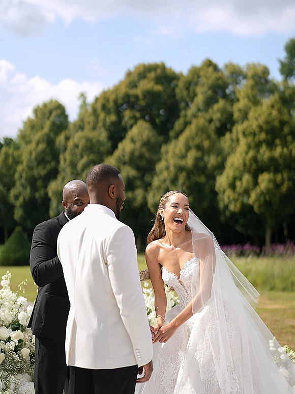 Ceremony moment at an outdoor wedding ceremony as bride laughs holding hands with groom, long veil and white flowers on a grassy lawn