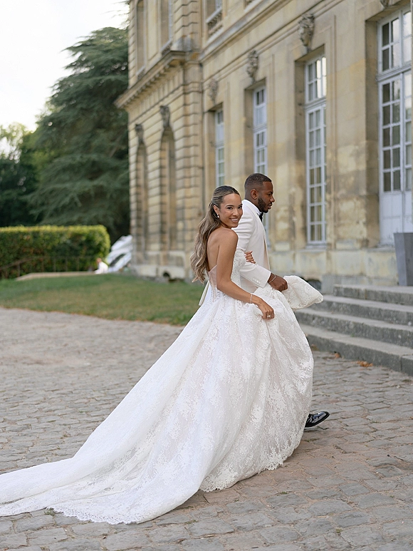 Couple portrait of bride looking back as they walk, her strapless lace gown with long train beside groom in white suit on stone steps