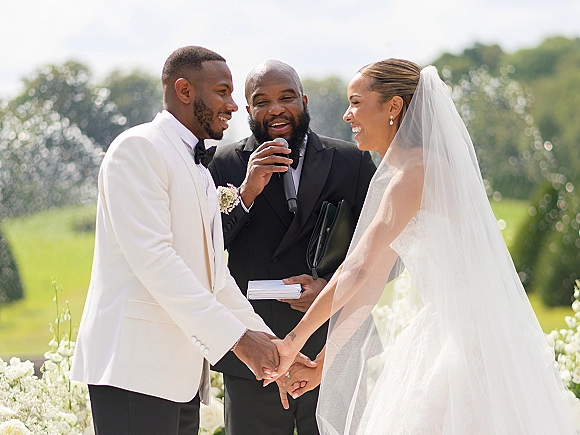 Wedding vows as the couple holds hands during an outdoor wedding ceremony, bride in veil and groom in tuxedo with officiant on mic outdoors