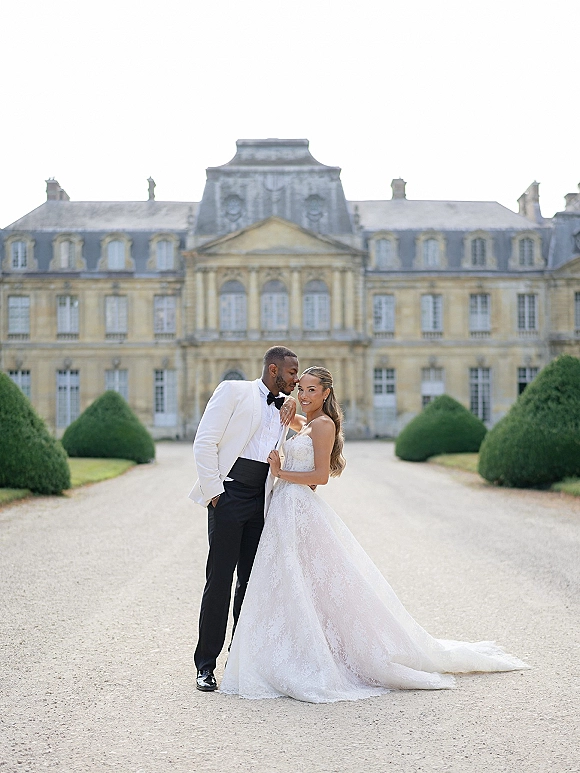Couple portrait of bride and groom pose on a gravel driveway, her strapless lace gown with long train before a grand estate facade