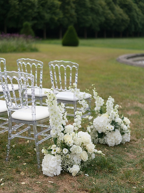Ceremony aisle decor with outdoor ceremony aisle flowers, clear acrylic chairs lined beside white rose and hydrangea clusters on a garden lawn