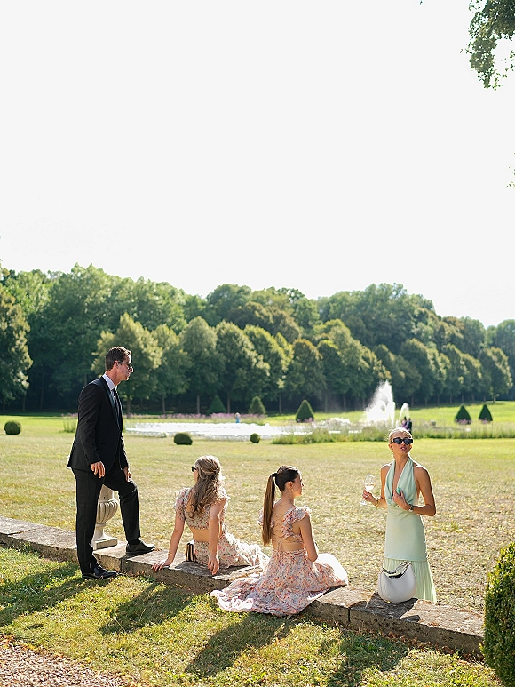 Wedding guests candid in wedding guest outfits chatting on a lawn by a fountain, some in sunglasses, holding wine near a stone ledge