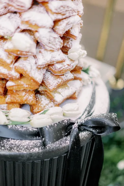 Beignet tower as a wedding dessert display, dusted with powdered sugar, set before lush greenery and a gold railing.