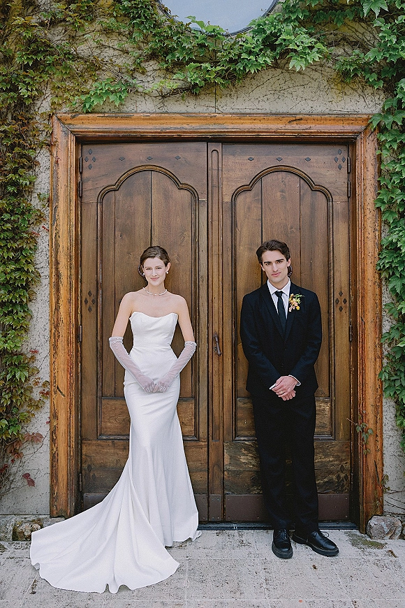 Couple portrait of bride in a strapless satin gown with gloves and pearl necklace beside groom in suit before ivy and wood doors