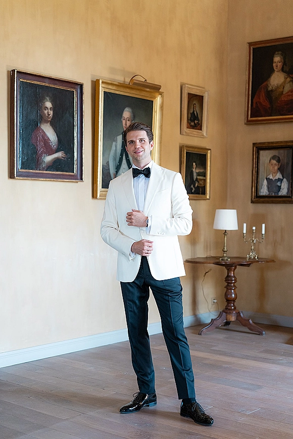 Groom portrait of a man in a white tuxedo with black bow tie, standing in a classic room with framed paintings and a table lamp