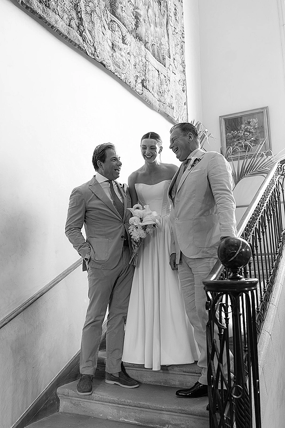 Wedding portrait of bride with parents photo on an indoor staircase, bride in strapless dress holding bouquet beside men in beige suits and boutonniere