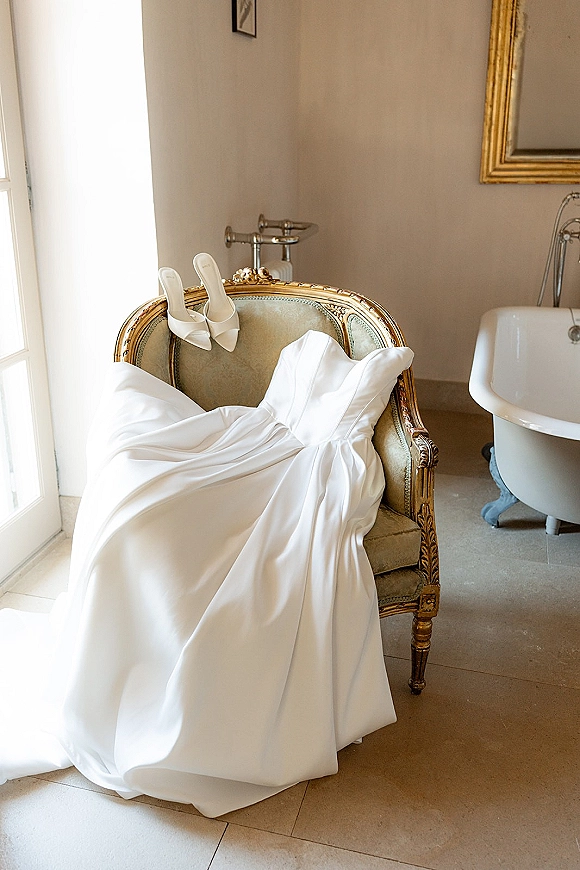 Wedding dress draped over a vintage chair with bridal heels below, lit by window light near a bathtub and wall mirror on tiled floor