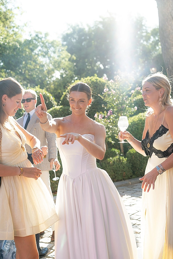 Wedding reception moment as the bride dances and points while laughing with friends holding wine glasses on a sunlit garden patio