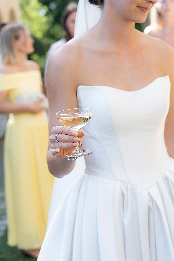 Wedding dress close-up of a strapless wedding dress with veil, bracelet, ring, and coupe glass at an outdoor cocktail hour with guests behind