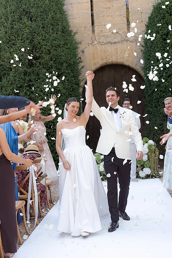 Wedding recessional rose petal toss as bride and groom walk the aisle runner, veil flowing, guests cheering in a stone-arch courtyard