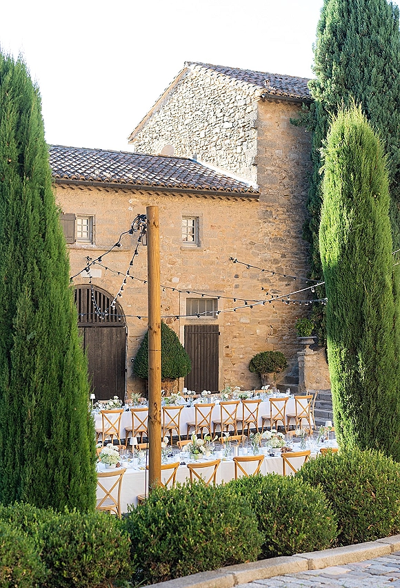 Outdoor reception setup in a courtyard wedding reception with long banquet tables, white linens, cross-back chairs, and string lights by a stone building