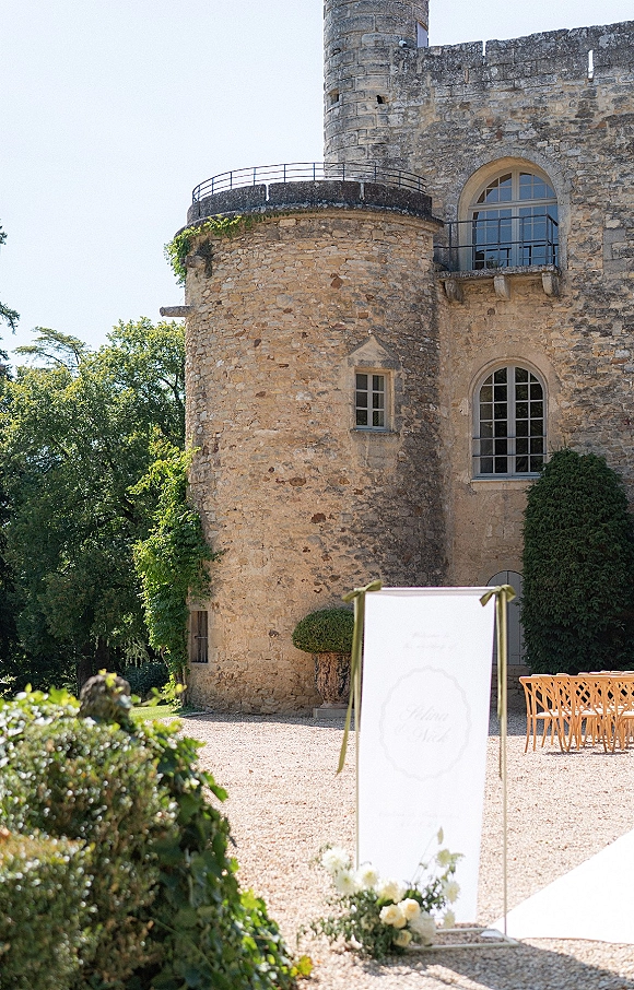 Wedding welcome sign in a gold frame with ribbon bows and a small floral arrangement, set in a sunlit stone castle courtyard