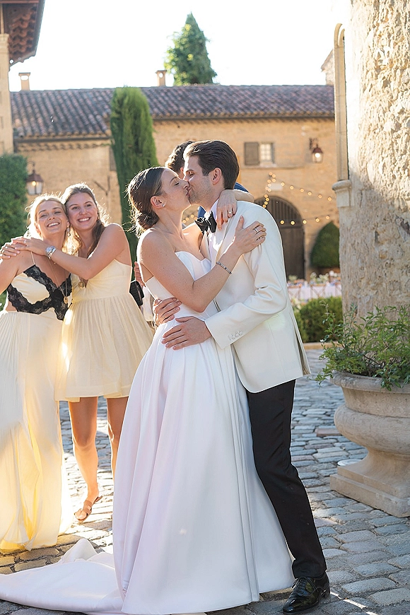Wedding kiss as bride in a strapless dress with train and groom in a white tuxedo jacket embrace in a stone courtyard with string lights