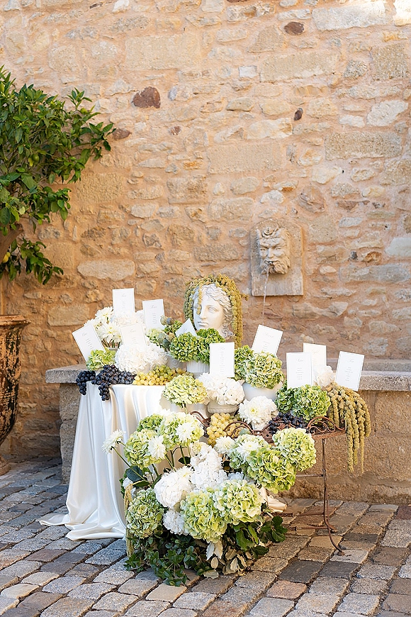 Wedding escort card display with hydrangeas, escort cards, grapes and greenery on a draped table beside a stone wall in a cobblestone courtyard