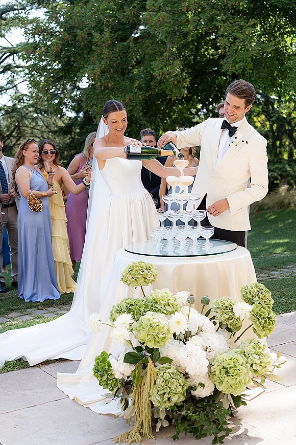 Champagne tower moment as bride and groom pour bubbly into coupe glasses at a garden table, veil flowing beside hydrangeas and guests