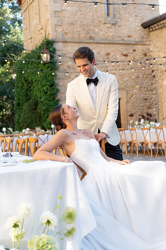 Couple portrait of bride and groom laughing at a reception table under string lights in an ivy-lined stone courtyard, bow tie and strapless dress