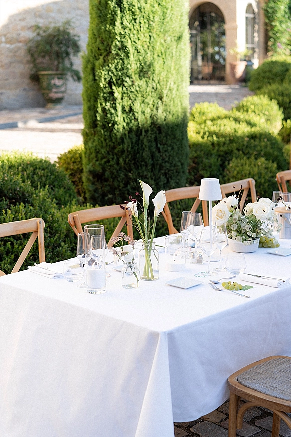 Reception tablescape with an outdoor reception table set in white linen, candles, lamps, bud vases, and grapes on a cobblestone patio by a stone archway