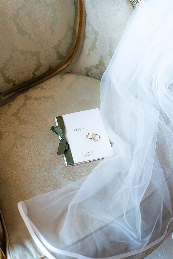 Wedding invitation suite with rings atop an envelope tied with a sage green ribbon bow, arranged on a vintage upholstered chair with veil
