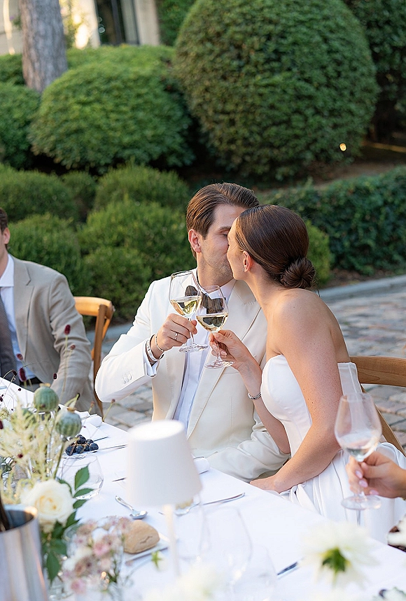 Wedding reception toast as newlyweds clinking glasses at a white tablecloth table with floral centerpiece on a stone patio with garden hedges behind