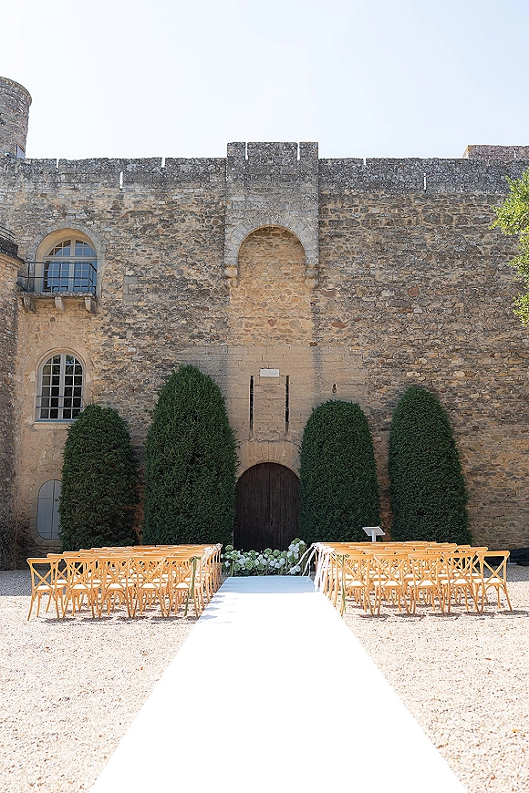 Ceremony aisle design with a white aisle runner leading past cross back chairs to a lectern, set in a gravel courtyard by stone castle walls