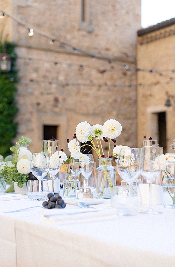 Reception tablescape with wedding table centerpiece of white floral clusters, bud vases, taper candles and grapes under string lights in a stone courtyard