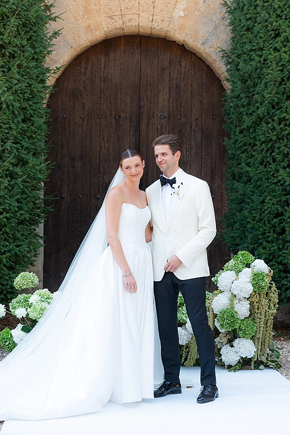 Couple portrait of bride in a strapless wedding dress and veil with groom in a white tuxedo jacket by a stone arch doorway outdoors.