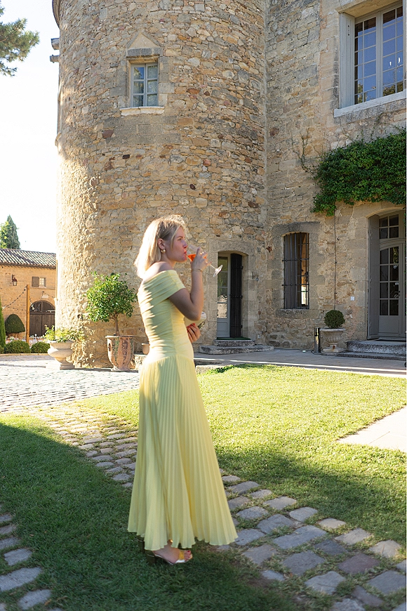 Wedding guest portrait in a yellow off-shoulder dress holding a cocktail in a stone castle courtyard with ivy and arched doorway