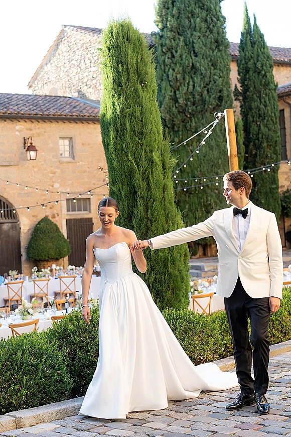 Couple portrait of bride and groom holding hands, her long train trailing on cobblestones in a stone courtyard lit by string lights.