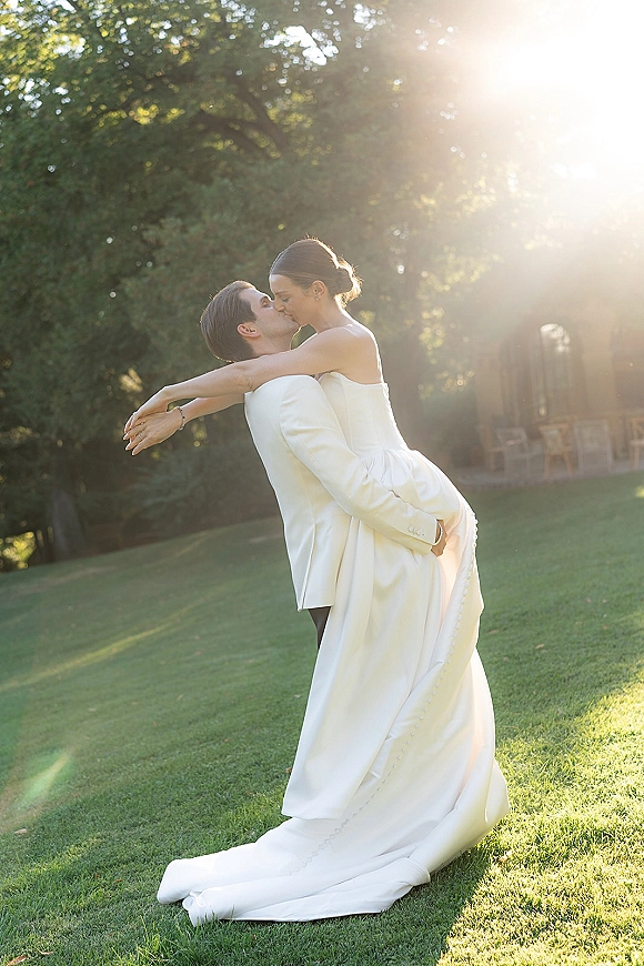 Wedding kiss as groom lifts bride in a strapless gown with long train, backlit by sun flare on a garden lawn with chairs