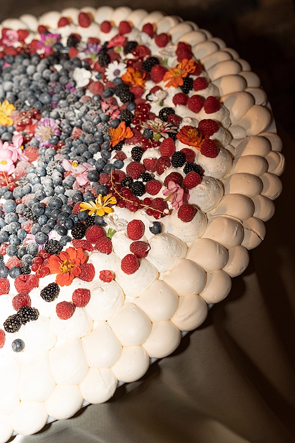 Wedding cake with white frosting and piped dollops, topped with mixed berries and edible flowers, dusted with powdered sugar on a dark backdrop
