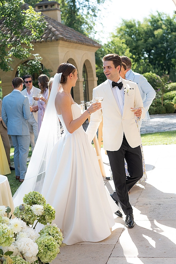 Wedding cocktail hour as bride and groom toasting with coupe champagne glasses, holding hands on a garden stone patio by an arched building