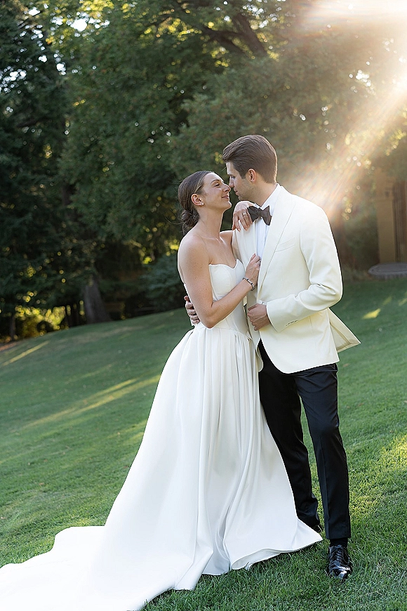 Couple portrait of bride and groom embrace nose to nose on a sunlit lawn with trees, sun flare, and a strapless full skirt gown