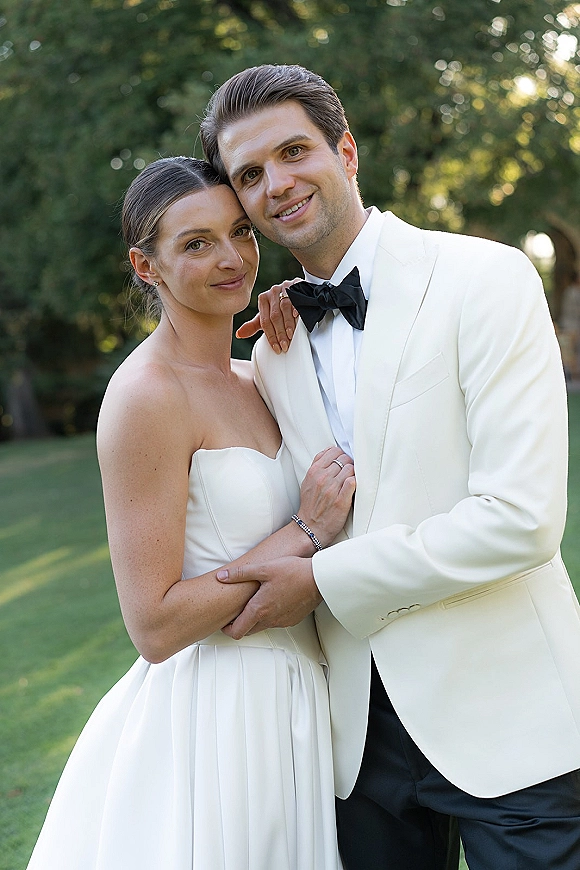 Couple portrait of bride in strapless wedding dress and groom in white tuxedo jacket with black bow tie, on sunlit garden lawn bokeh