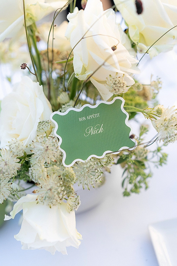 Wedding place card with a scalloped edge in green tucked into a white rose and Queen Anne’s lace centerpiece on a white reception table
